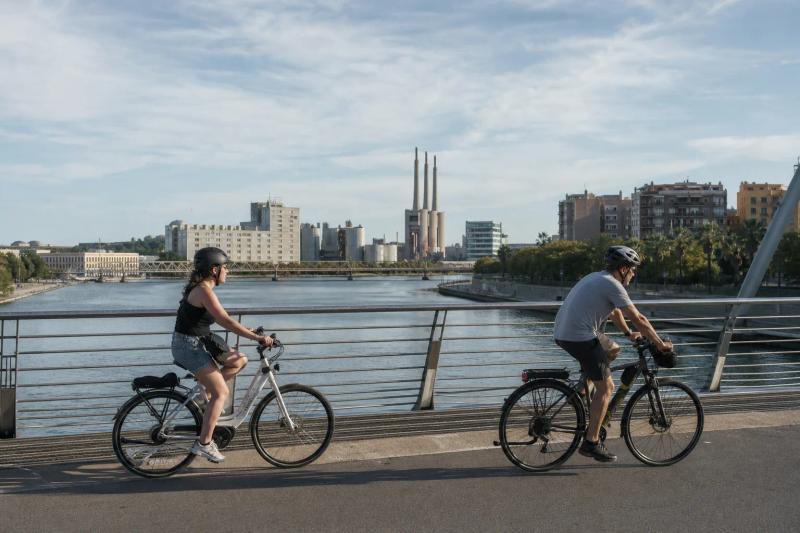 Cyclists crossing a river bridge in Barcelona with city buildings behind